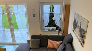 Ein Wohnzimmer mit einer Eckcouch, einem Couchtisch, einem kleinen Beistelltisch, einem Fenster mit Blick auf eine grüne Rasenfläche und einem gerahmten Bild an der Wand.