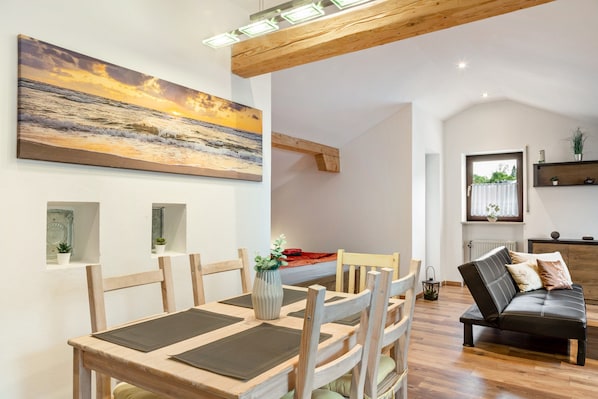 A dining area with a wooden table and chairs, a painting of a sunset over the ocean, and a cozy seating area with a sofa and cushions.