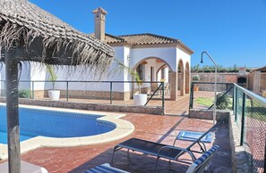A swimming pool with lounge chairs, a thatched roof structure, and a patio area with a brick building and arches. - Villa Ureba        . (Conil de la Frontera)