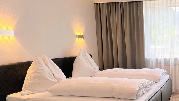 Habitación de hotel con una cama, dos lámparas de pared, una ventana con cortinas y un accesorio de iluminación en el techo.