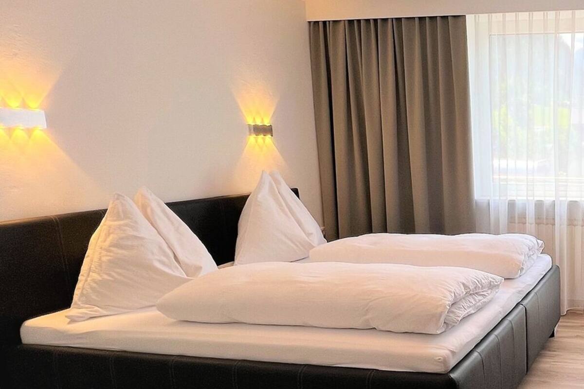 A hotel room with a bed, two wall-mounted lights, a window with curtains, and a ceiling light fixture.