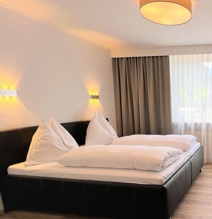 A hotel room with a bed, two wall-mounted lights, a window with curtains, and a ceiling light fixture. - Appartements by Hofer (Kaprun)