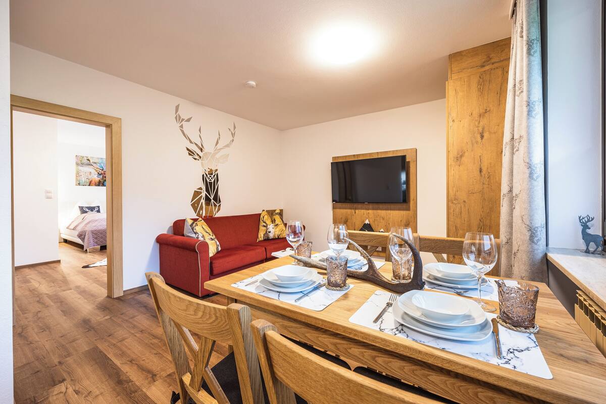 A dining area with a wooden table set for four, a red sofa, and a television mounted on the wall.