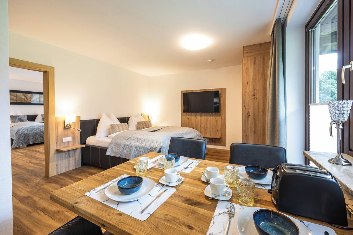 A modern hotel room with a wooden dining table set for breakfast, a bed with a TV, and a large window with curtains.