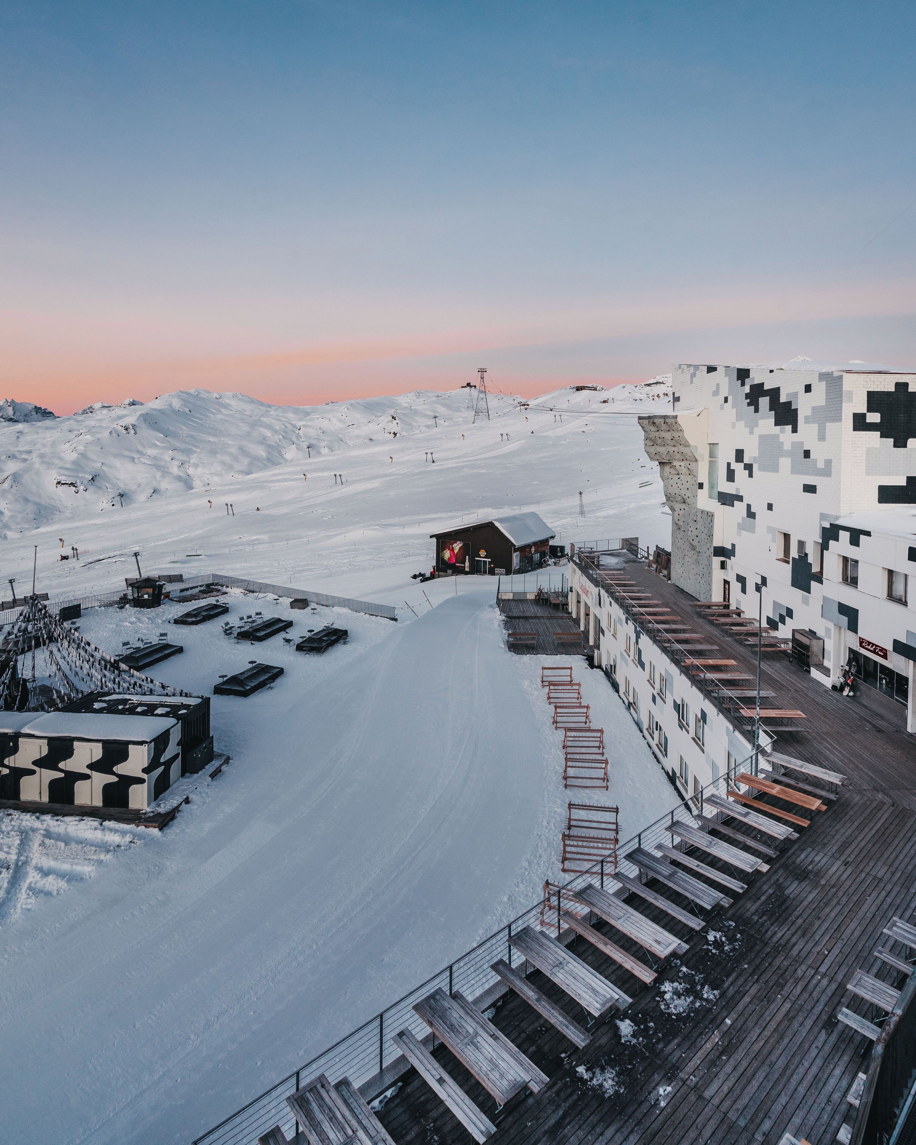Une station de ski enneigée, dotée d’une terrasse en bois et d’un bâtiment à la façade pixelisée.