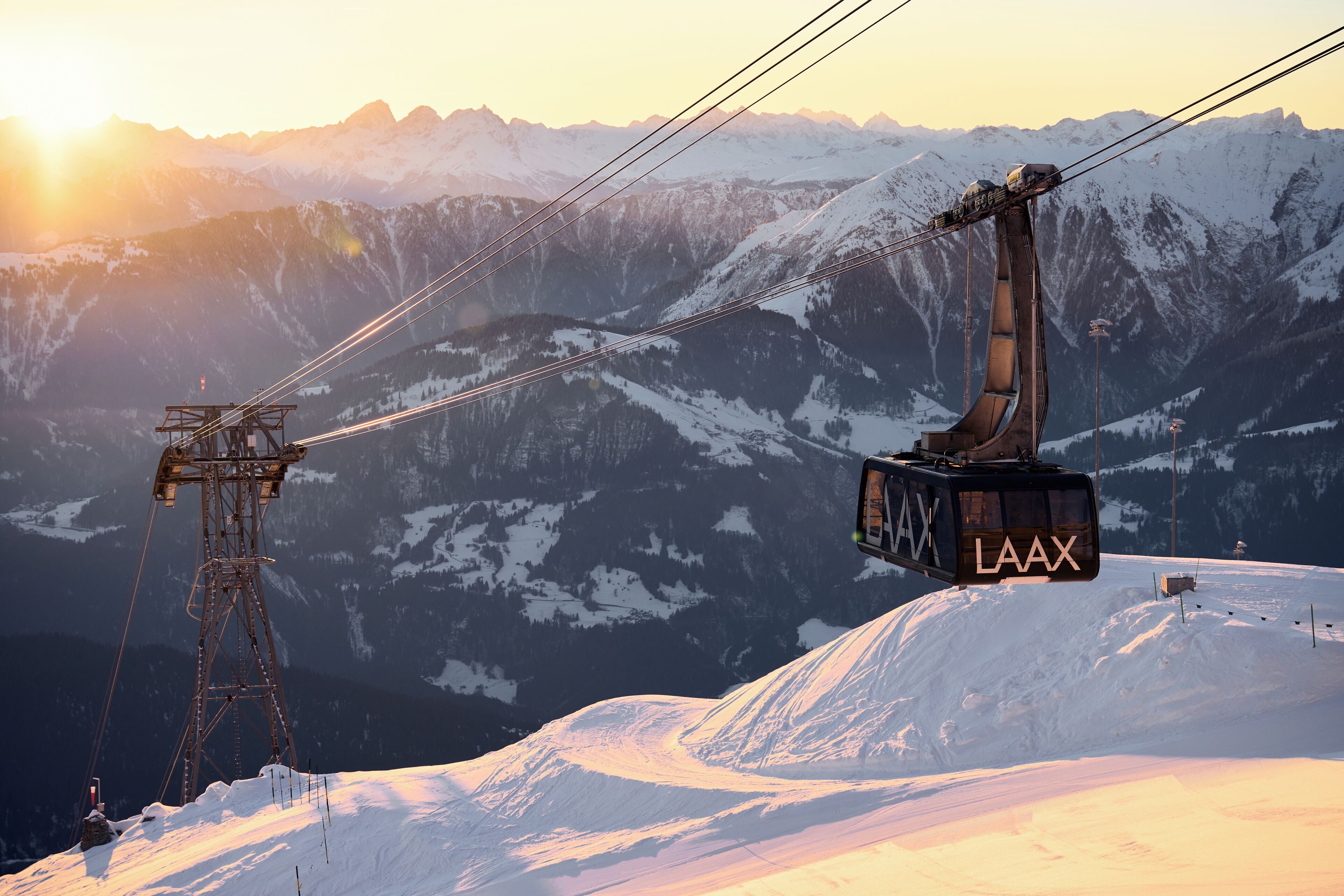 Sistema de teleférico con un vagón visible marcado con la etiqueta "LAAX" suspendido entre dos torres, con montañas nevadas y un sol poniente al fondo.