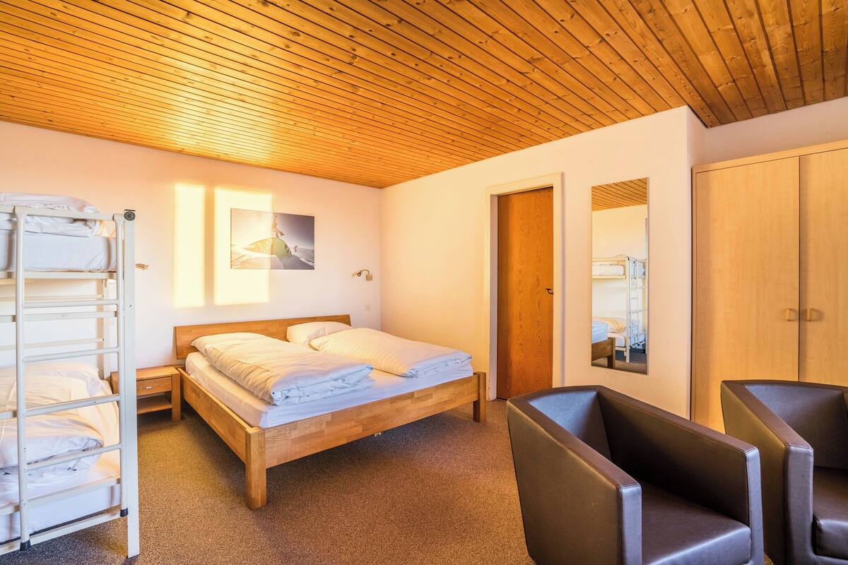 A bedroom with a wooden ceiling, a bed with white linens, a wooden wardrobe, and a seating area with two chairs.