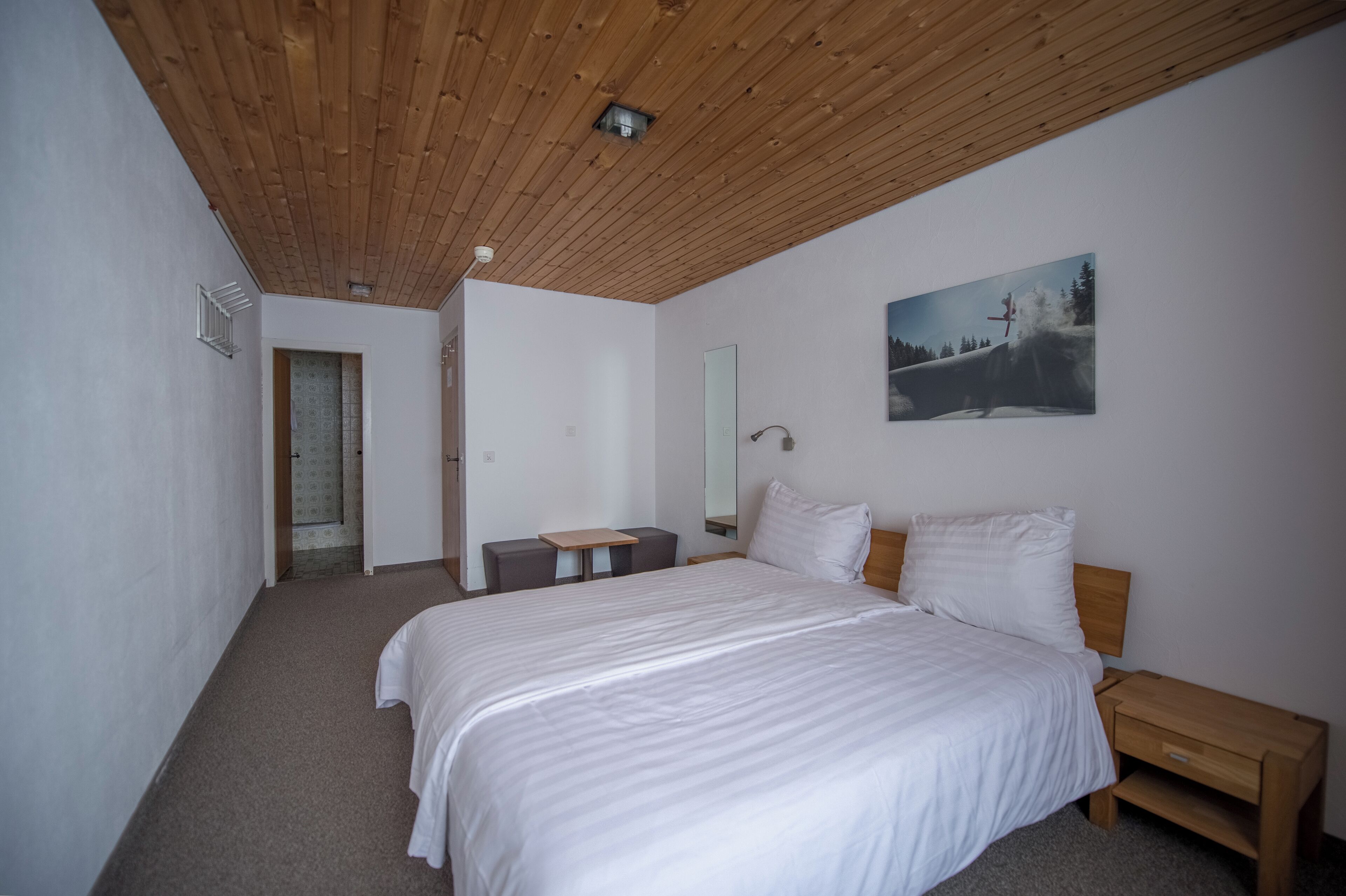 A bedroom with a wooden ceiling, a bed with white bedding, a bedside table, a nightstand, a small desk, and a painting on the wall.
