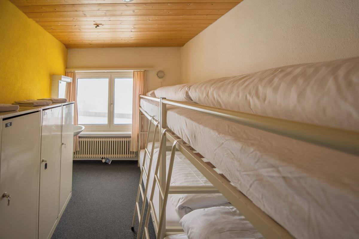 A dormitory room with bunk beds, a radiator, a window with curtains, and a yellow wall.