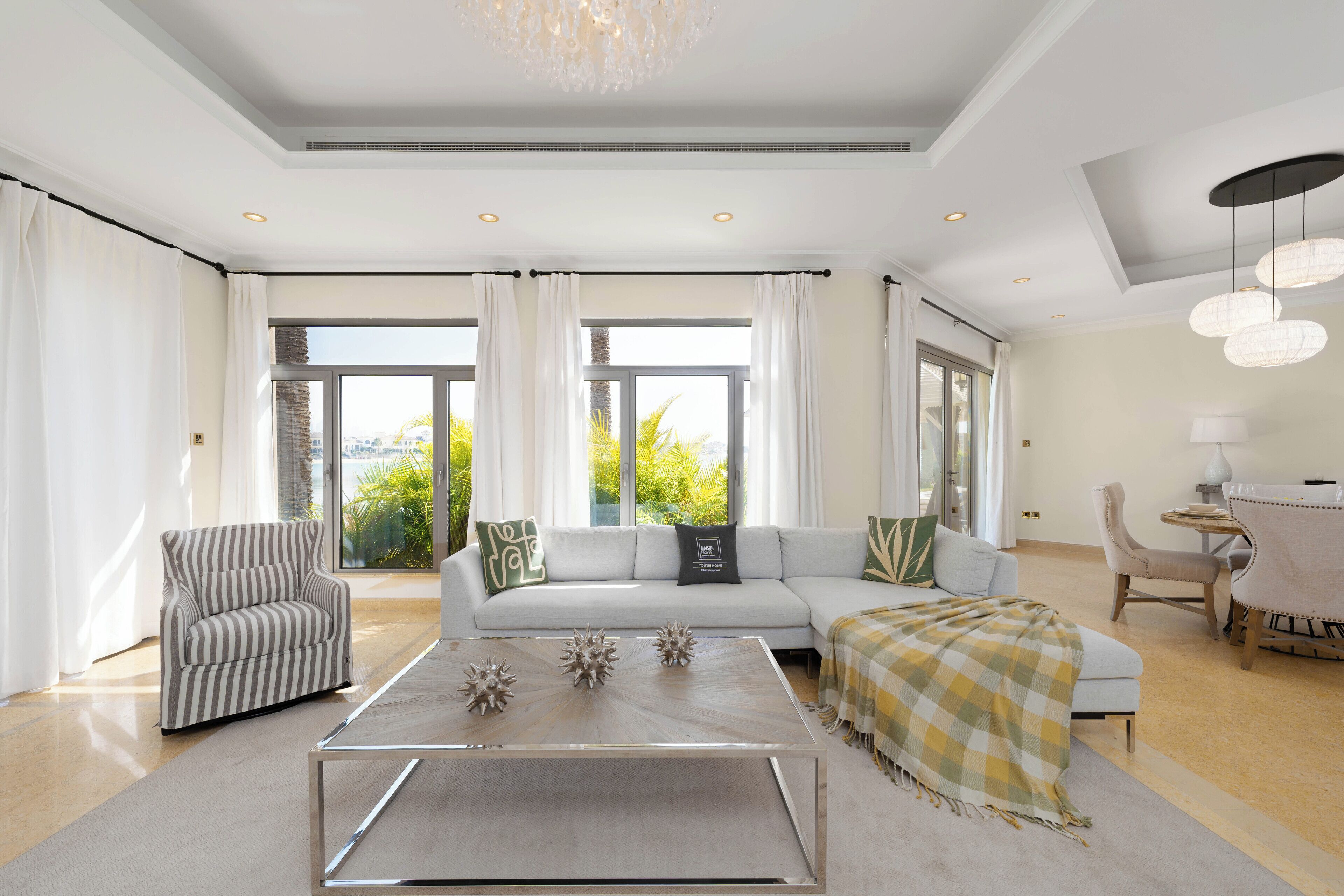 A spacious living room with a sofa, armchair, and coffee table, large windows with curtains, and a chandelier.