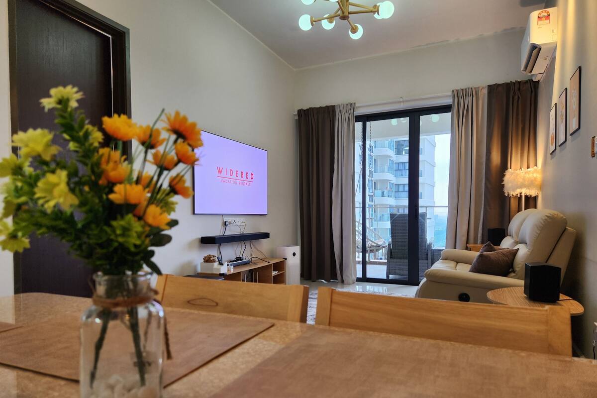 Una sala de estar moderna con un televisor mostrando “WIDEBED Vacation Rentals”, un comedor con un jarrón de flores y vistas a un balcón con cortinas.