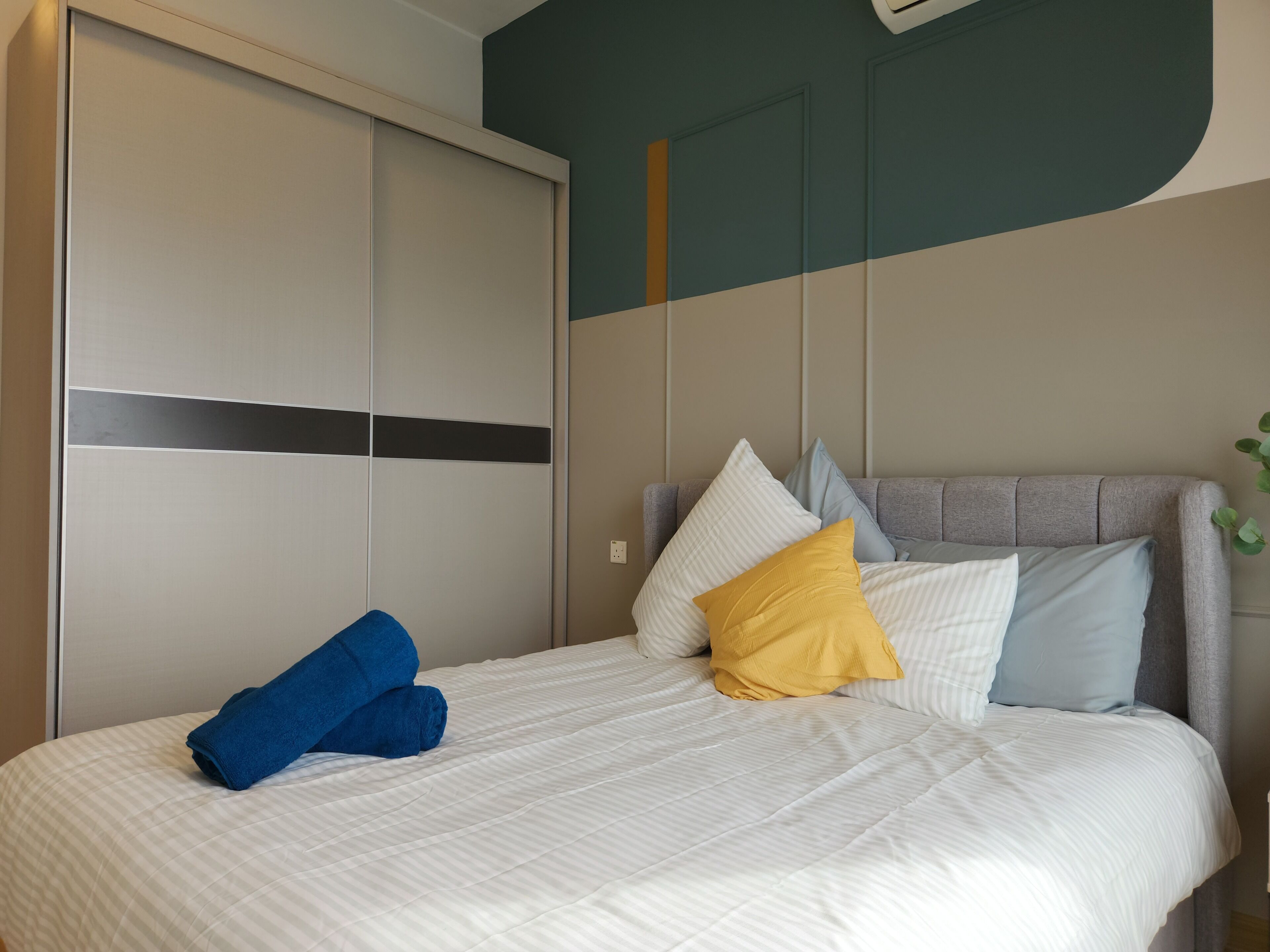 A neatly made bed with a white comforter and multiple pillows, a grey upholstered headboard, and a large wardrobe with sliding doors.