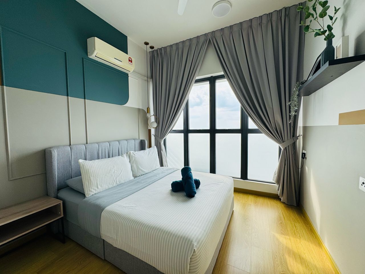A modern hotel room with a bed, a bedside table, a window with curtains, and an air conditioning unit.