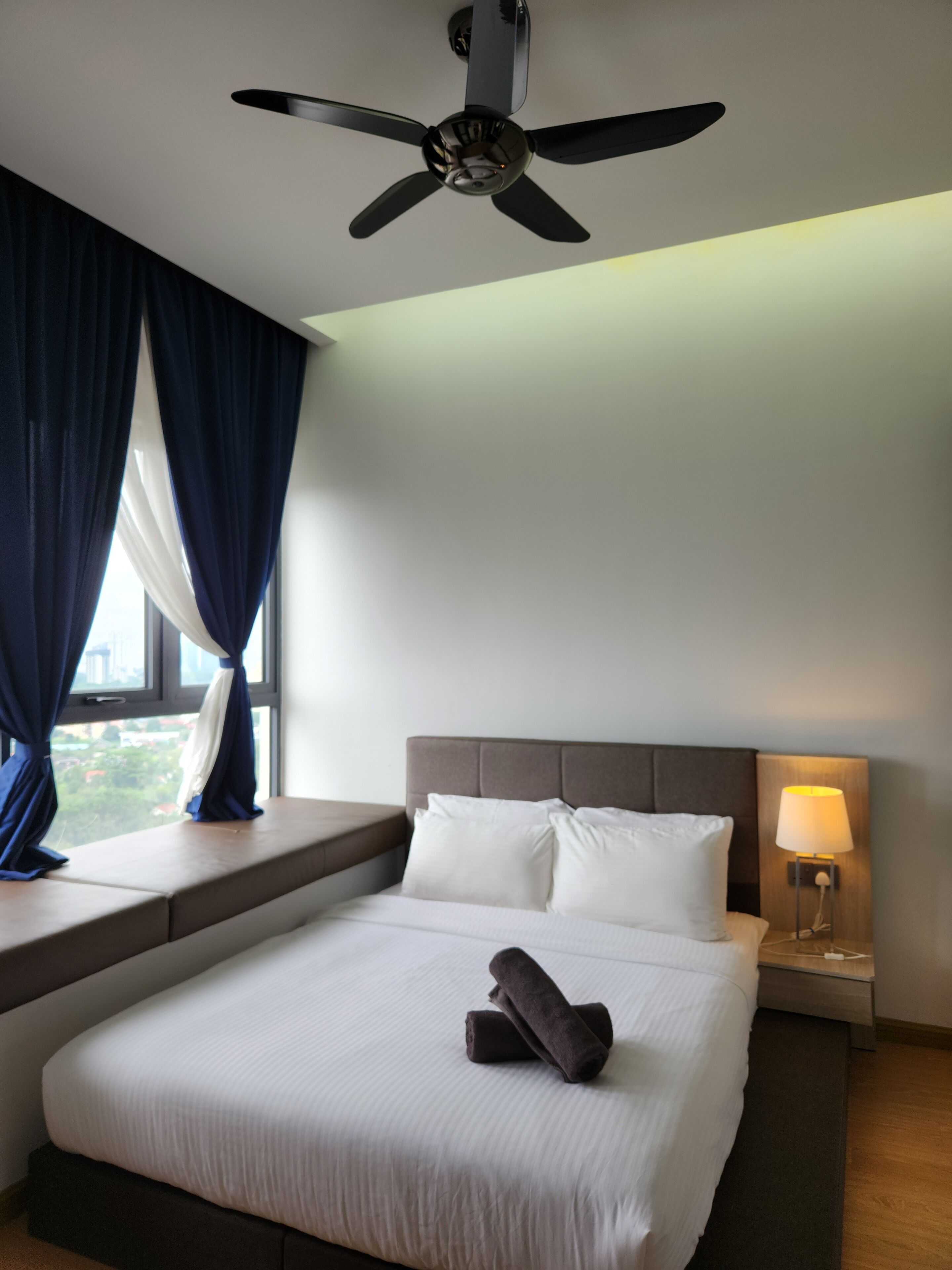 Een hotelkamer met een bed, een plafondventilator, een nachtkastje met een lamp en een raam met gordijnen.