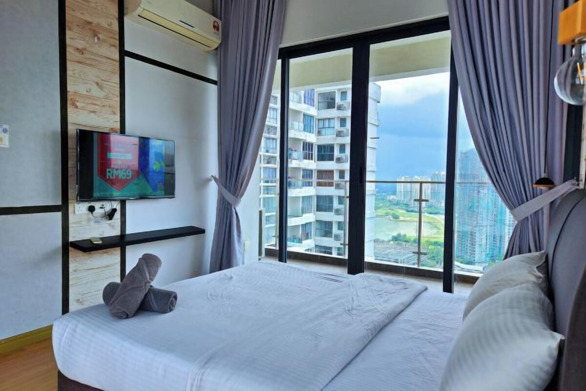 Habitación de hotel con cama, televisión, ventana con cortinas y vistas a edificios y al mar.