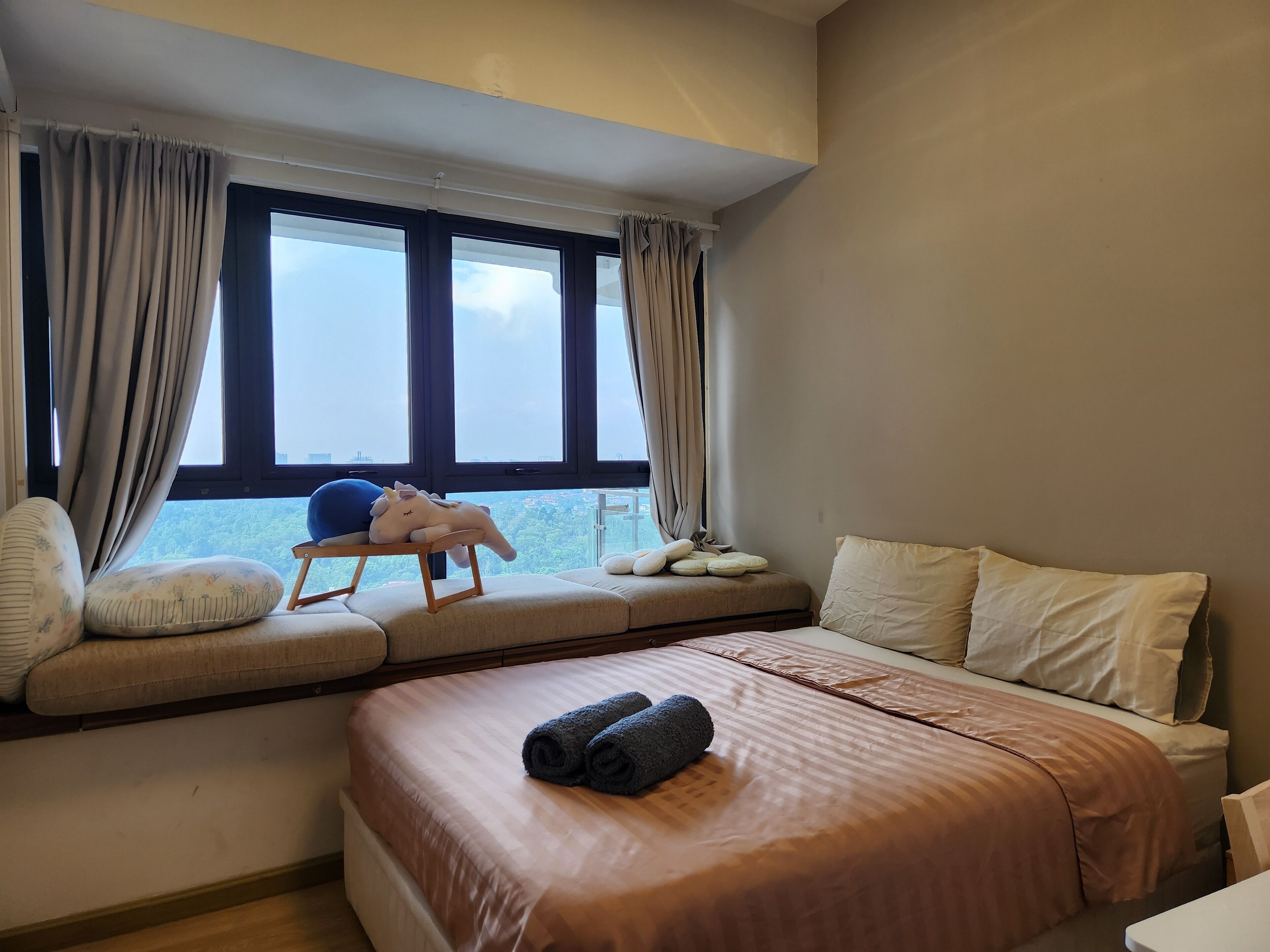 A bedroom with a large bed, a window seat with a plush toy, and a view of the cityscape.