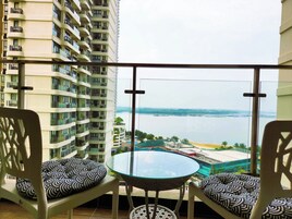 A balcony with two white chairs and a glass table, overlooking a body of water and a distant shoreline.