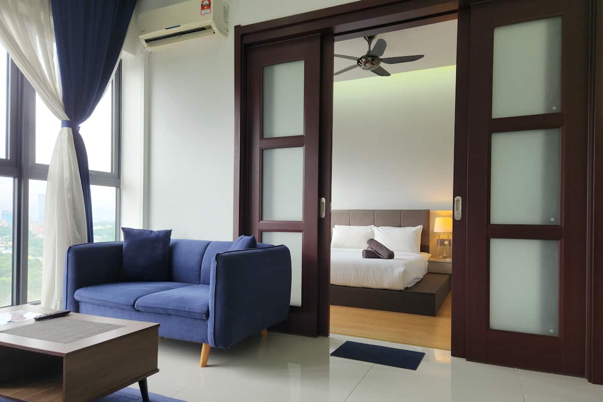 Habitación de hotel moderna con un sofá azul, una mesa de centro de madera, una cama con ropa de cama blanca, un ventilador de techo y vistas a la ciudad a través de grandes ventanales.