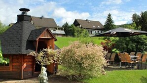 Una cabaña de madera rústica con chimenea, rodeada por un jardín cuidado y un área de estar al aire libre.