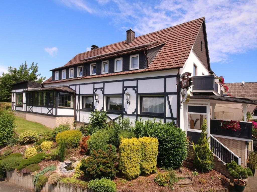 A traditional half-timbered building with a steeply pitched roof, multiple windows, and a well-maintained garden with various shrubs and a small pond.