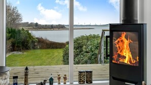 Een moderne woonkamer met een groot raam met uitzicht op een meer en windturbines, een open haard met een vlam en een houten terras met buitendecoratie.