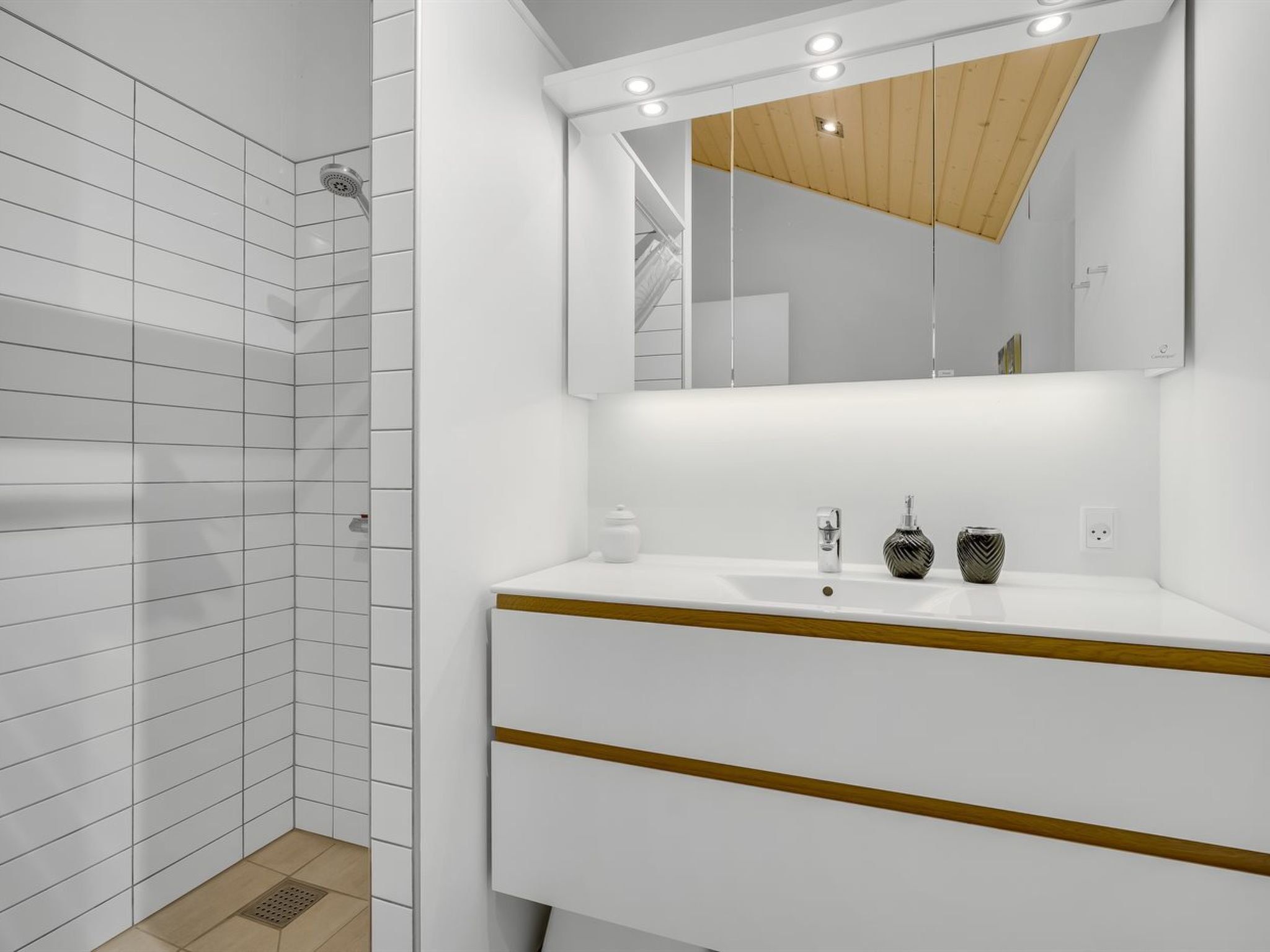 Ein modernes Badezimmer mit weißen Fliesen, einem Holzlagerfläschchen und einem Spiegel unter einer Holzbalkendecke.