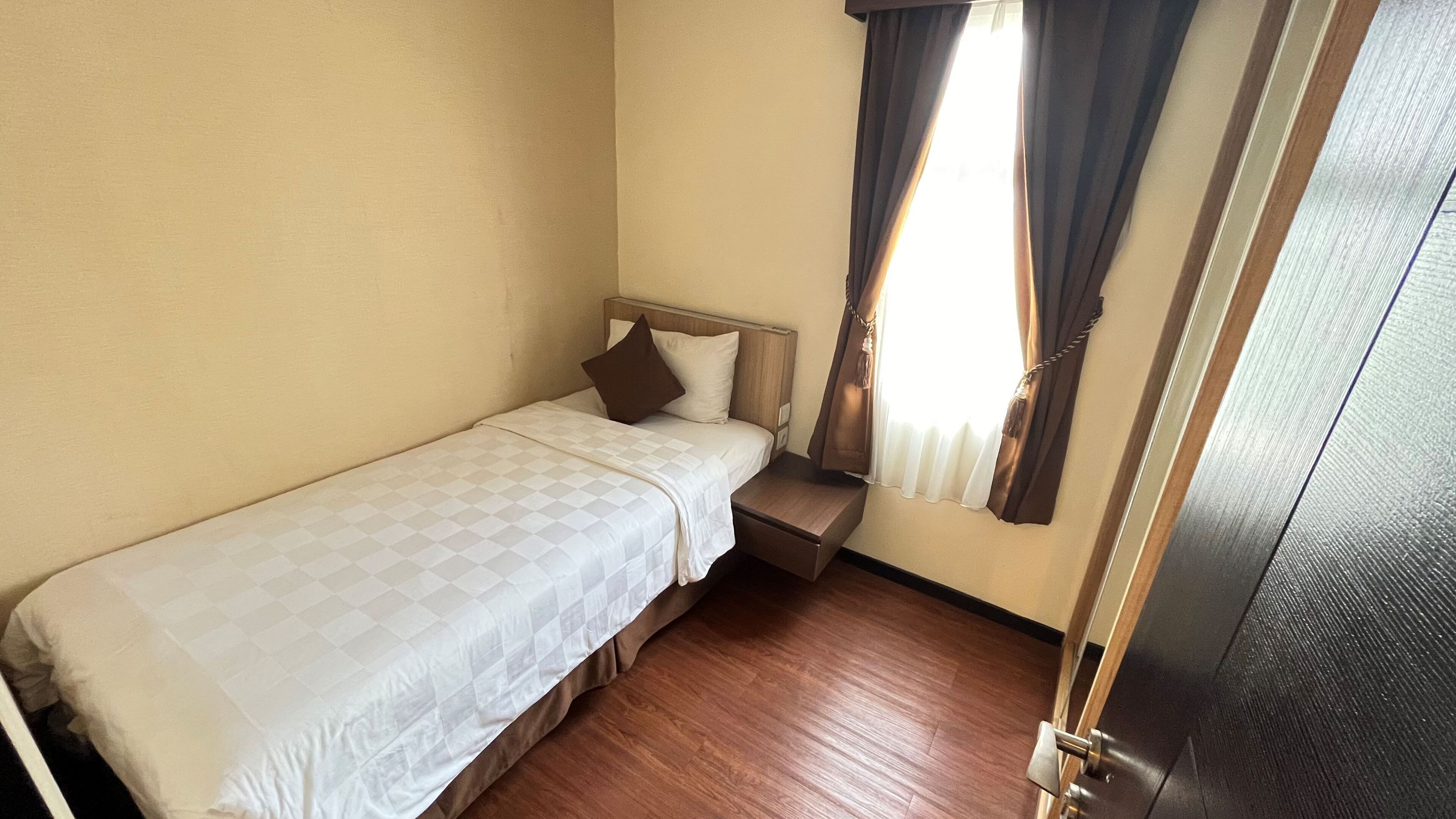 Una habitación pequeña con una cama individual, cabecera de madera, mesita de noche y ventana con cortinas.
