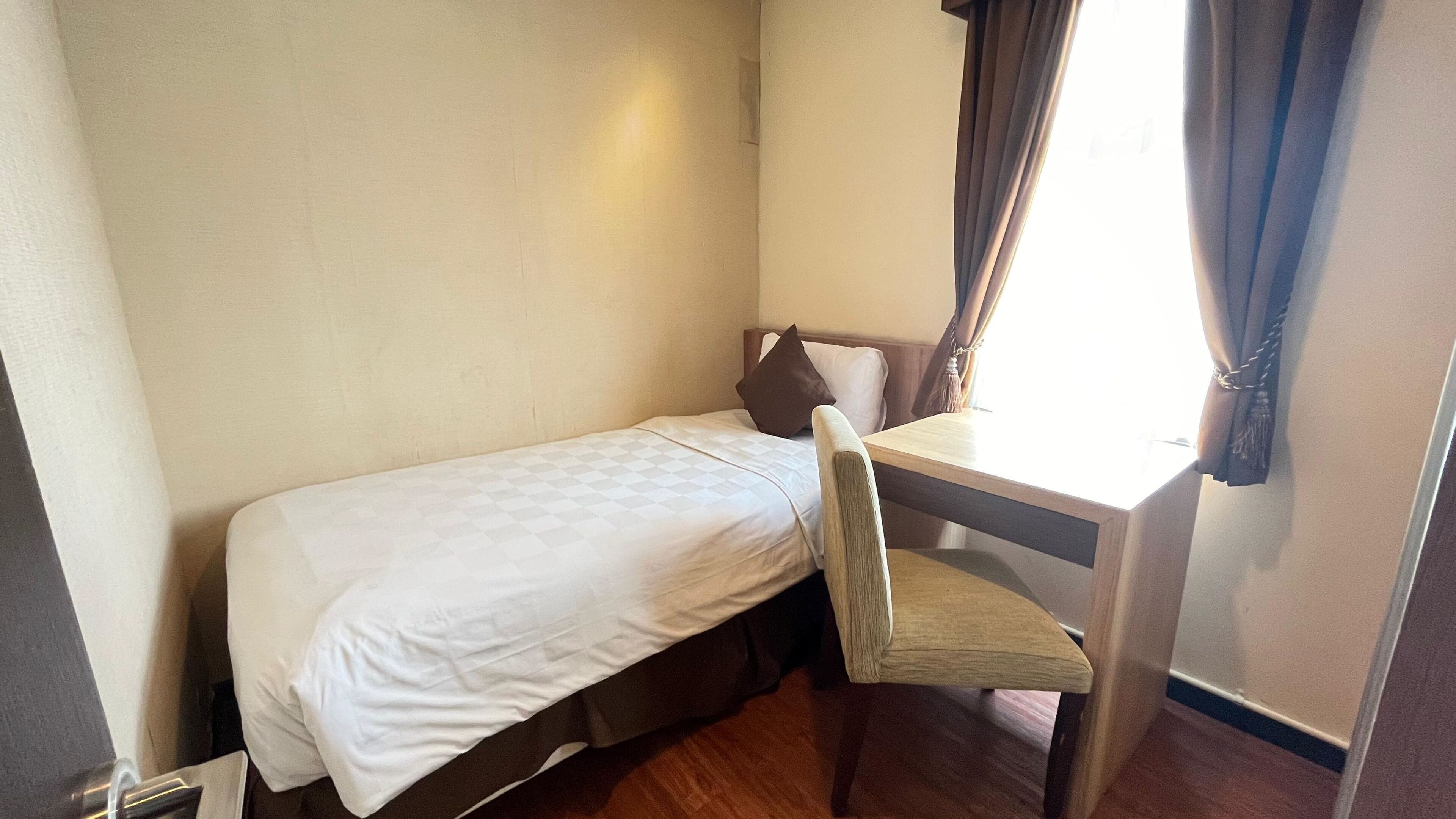 A small, single-bed room with a wooden desk and chair, a window with curtains, and a plain wall.