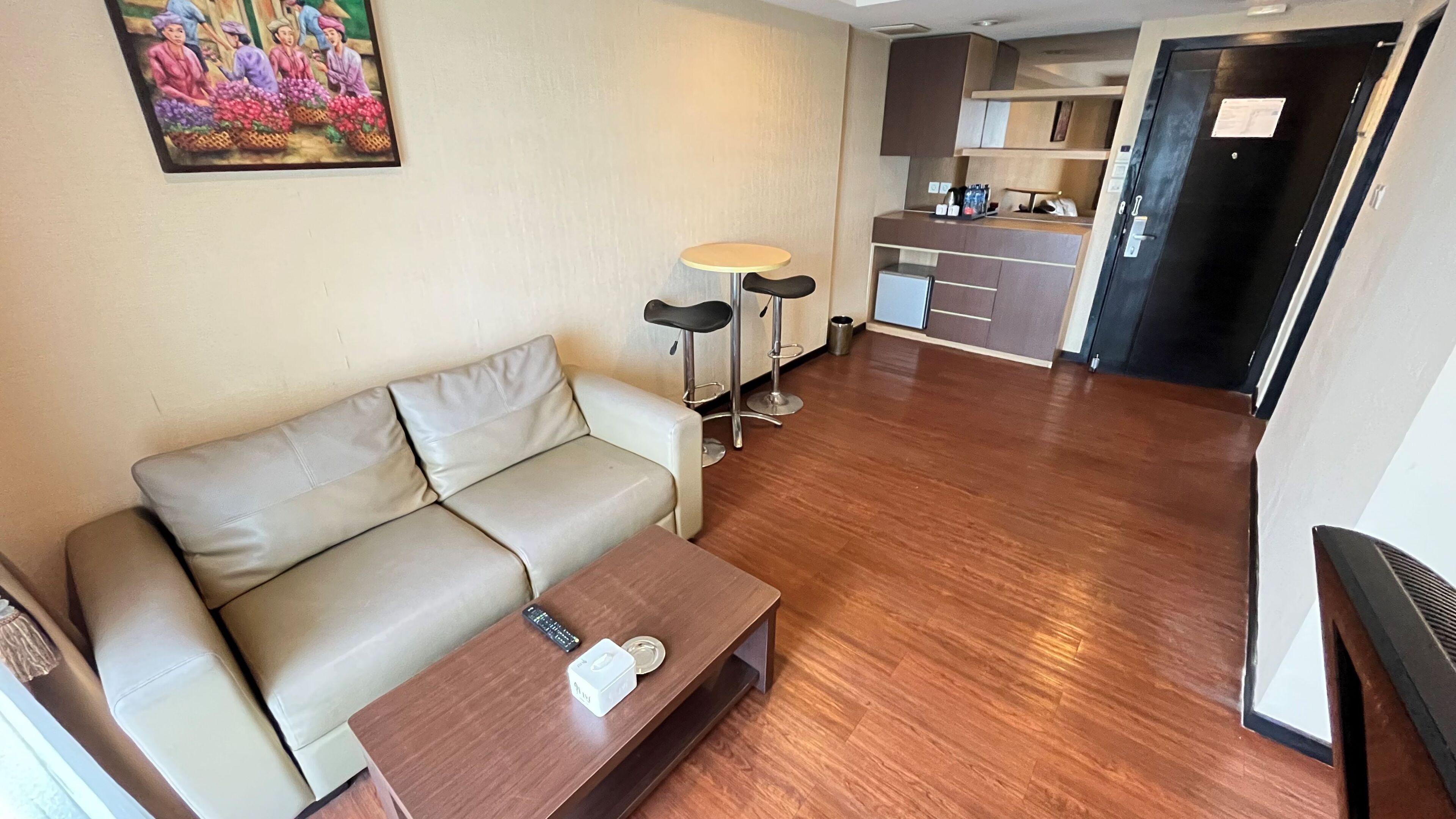 A modern living room with a sofa, a coffee table, and two bar stools. There is a kitchen area with cabinets and a refrigerator in the background.
