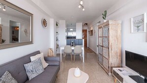 En moderne stue med en grå sofa, et sofabord og et fjernsyn. Der er et spiseområde med et bord og stole, og et køkken i baggrunden.