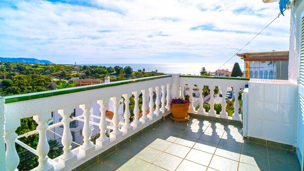 Un balcón con barandilla blanca, una planta en maceta y vistas de un paisaje costero.