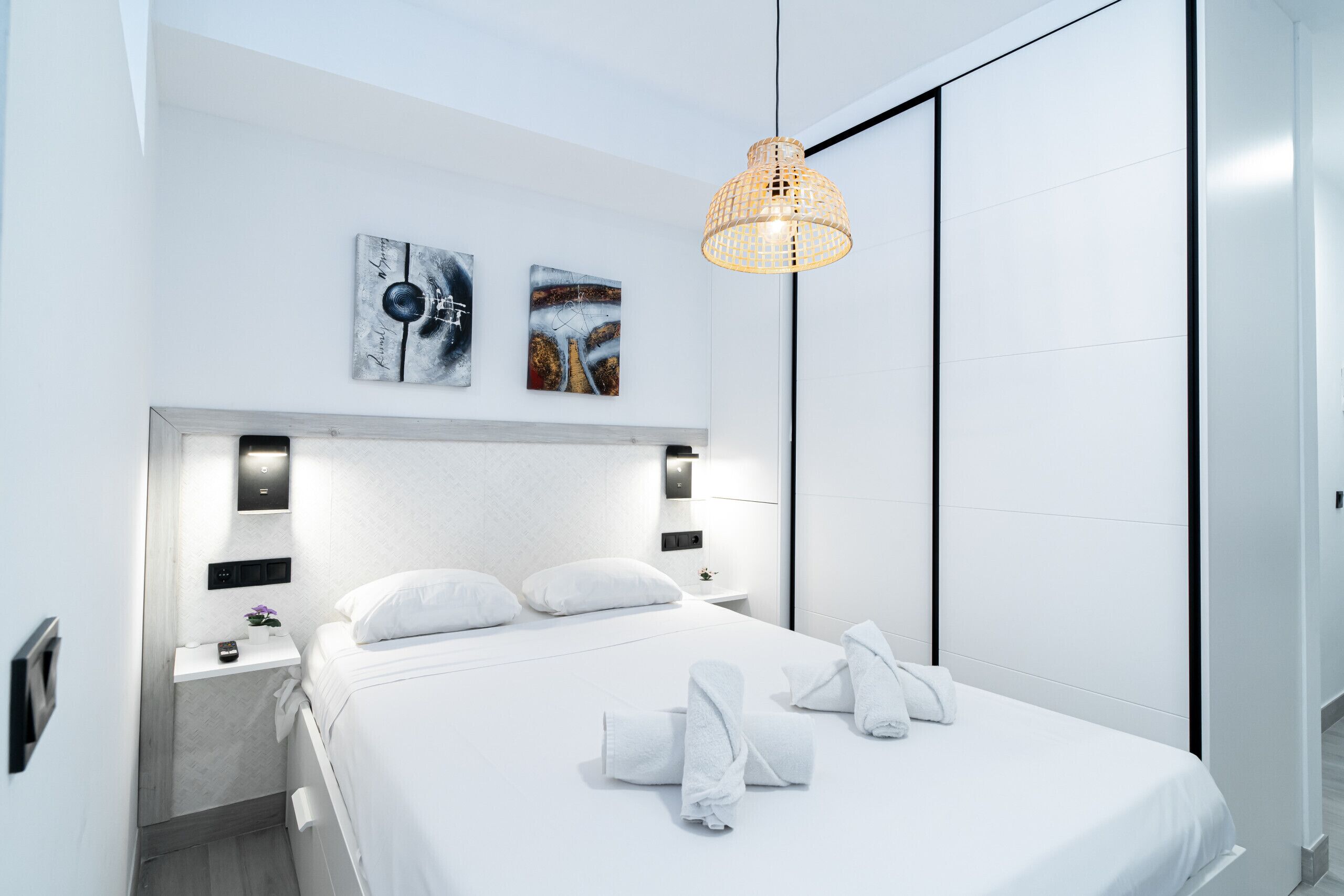 A modern hotel room with a large bed, white linens, a bedside table with a lamp, and artwork on the wall.