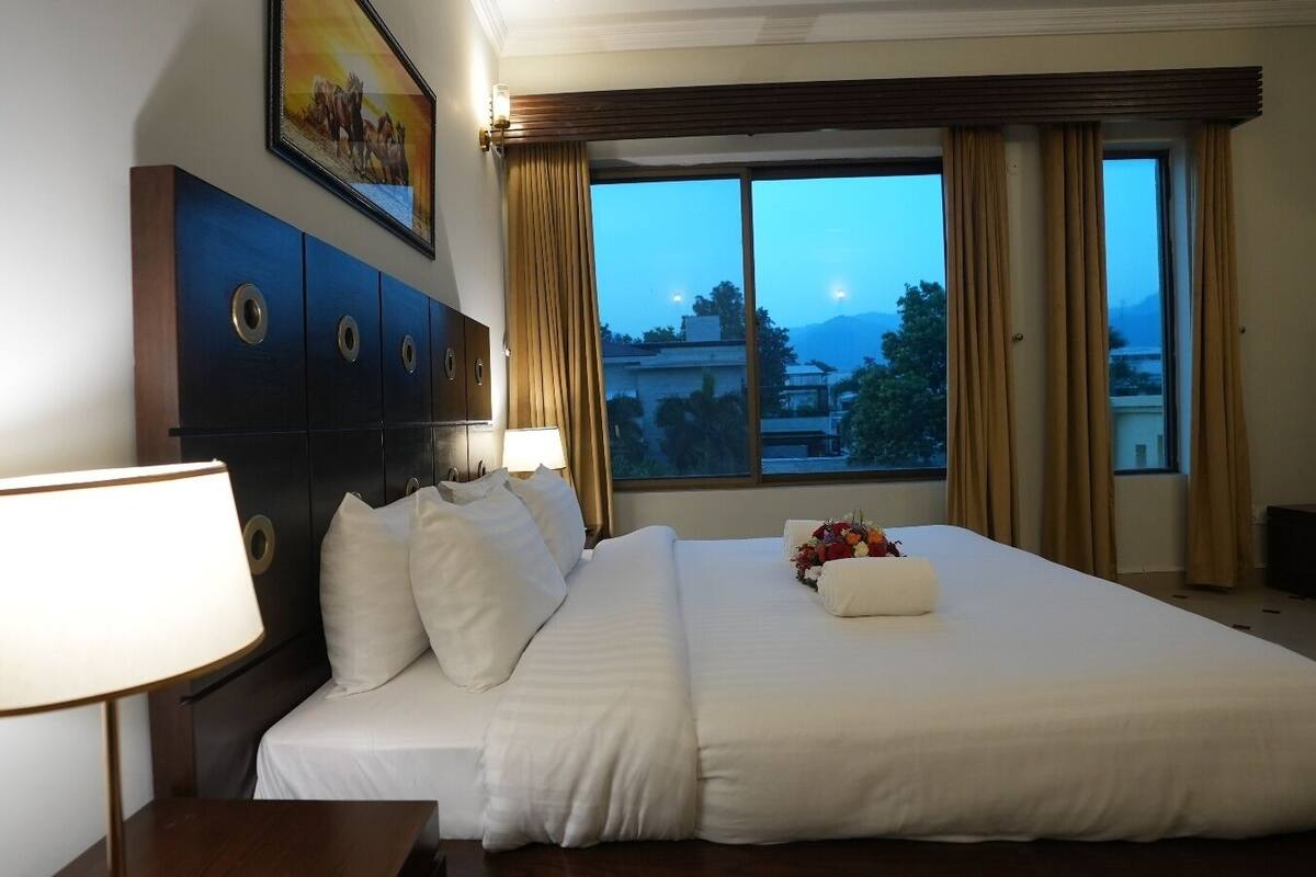 A hotel room with a large bed, a nightstand with a lamp, a wall-mounted TV, and a window with curtains.