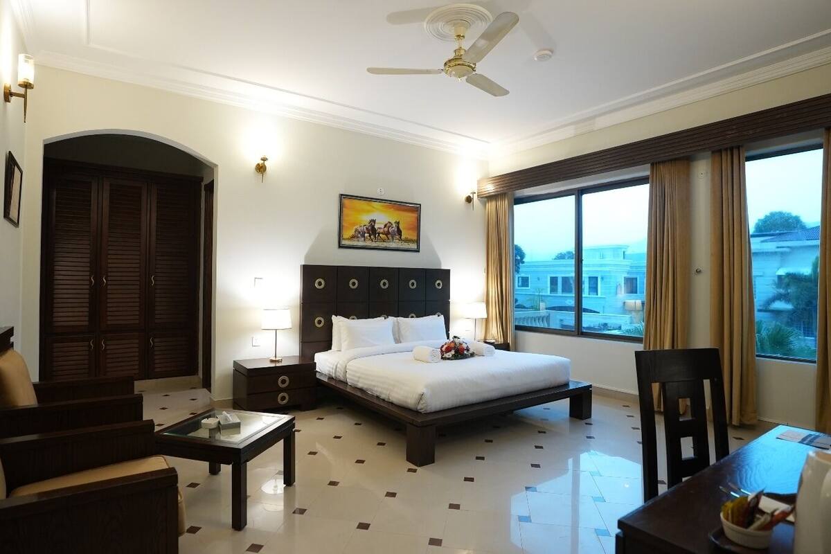 A hotel room with a bed, a desk, a chair, a nightstand, a ceiling fan, and a large window with curtains.