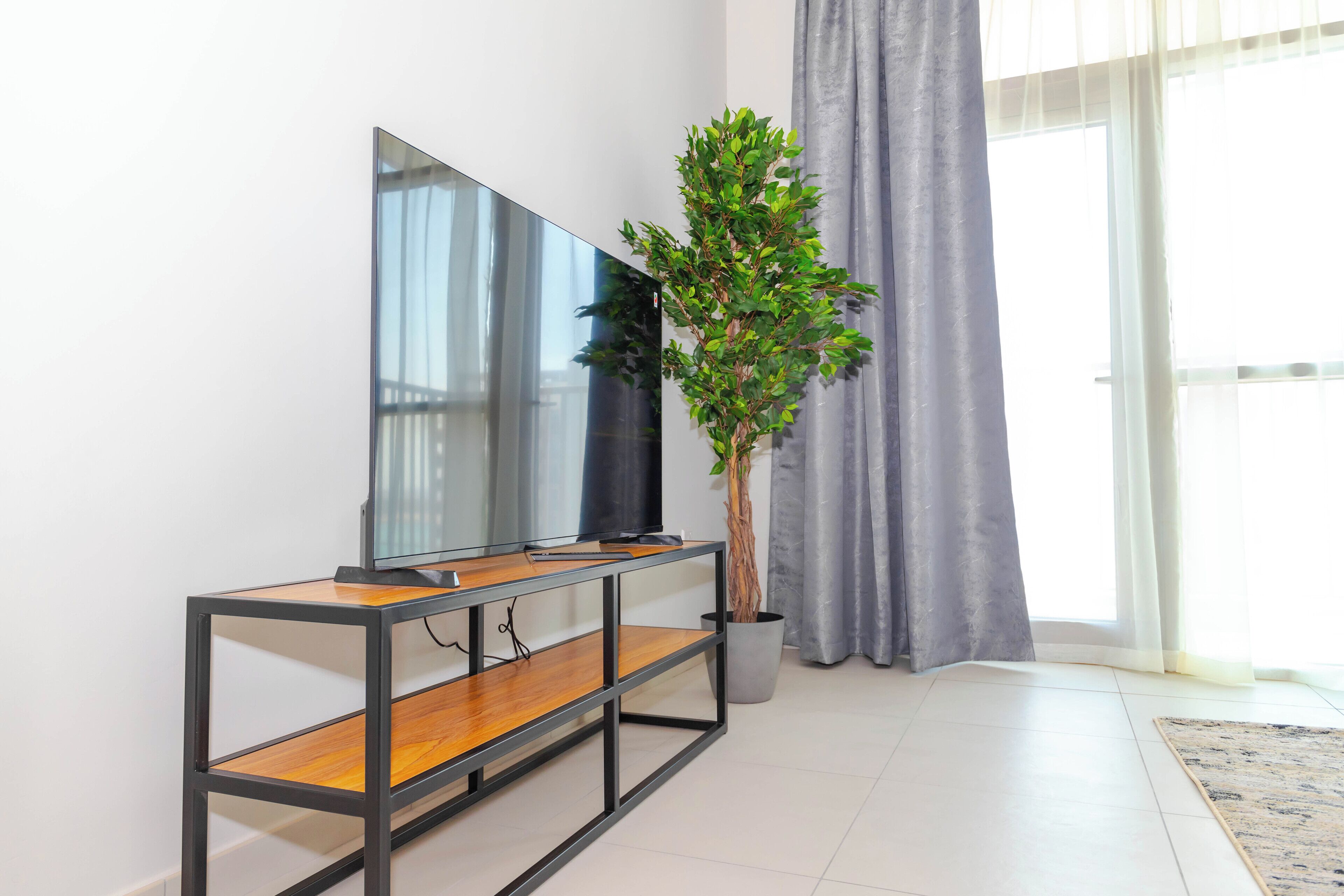 A modern living room with a flat-screen TV on a wooden and metal console table, a potted plant, and a large window with curtains.