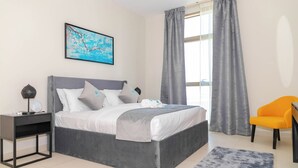 Ein modernes Schlafzimmer mit grauem Bett, weißer Bettwäsche, einem grauen Nachttisch mit Lampe, einem gerahmten Bild an der Wand und einem Fenster mit grauen Vorhängen.
