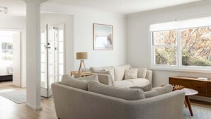 A modern living room with a beige sofa, a small wooden side table, and a large window with a view of trees.