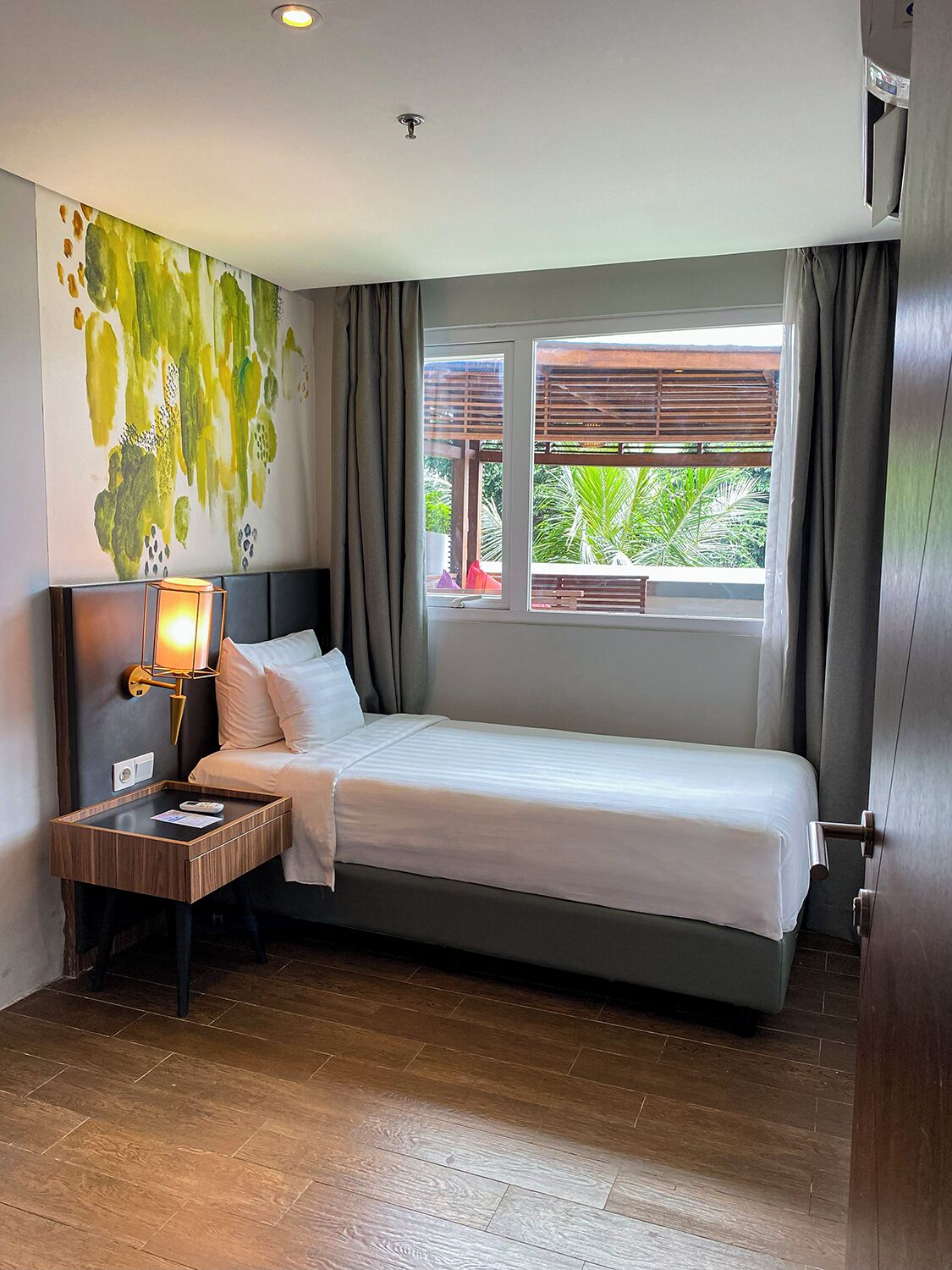 A hotel room with a bed, a nightstand, a lamp, a wall art piece, and a window with a view of greenery.