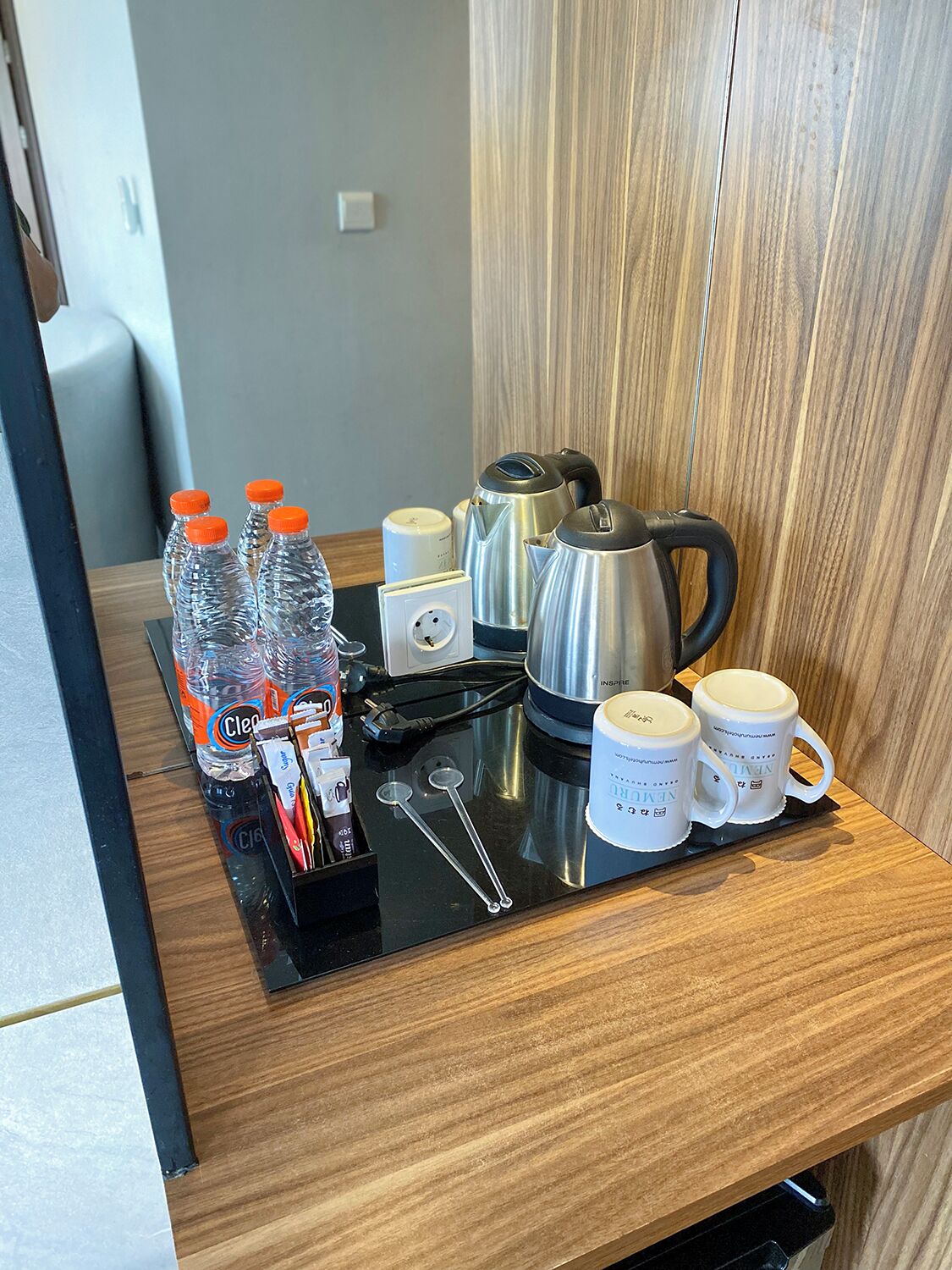 Une kitchenette dans une chambre d’hôtel, équipée d’un plan de travail en bois, d’une bouilloire, d’une machine à café et de plusieurs bouteilles d’eau.