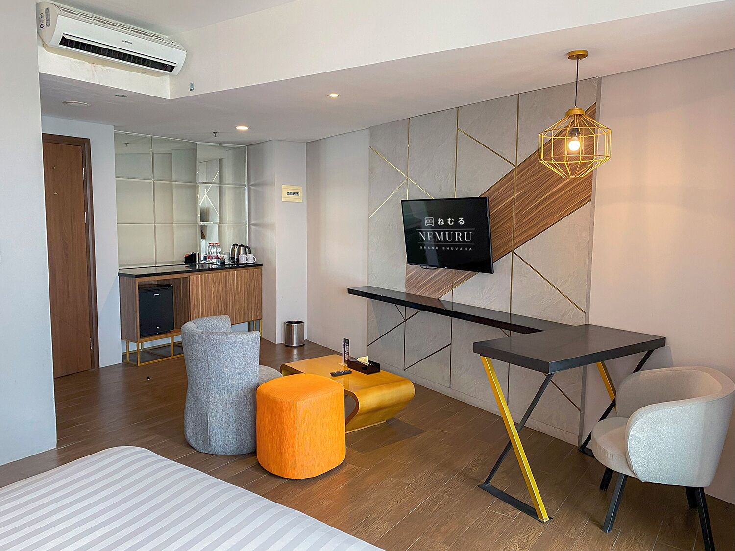 A modern hotel room with a flat-screen TV, a wooden accent wall, a small kitchenette, and a seating area with ottomans and a chair.