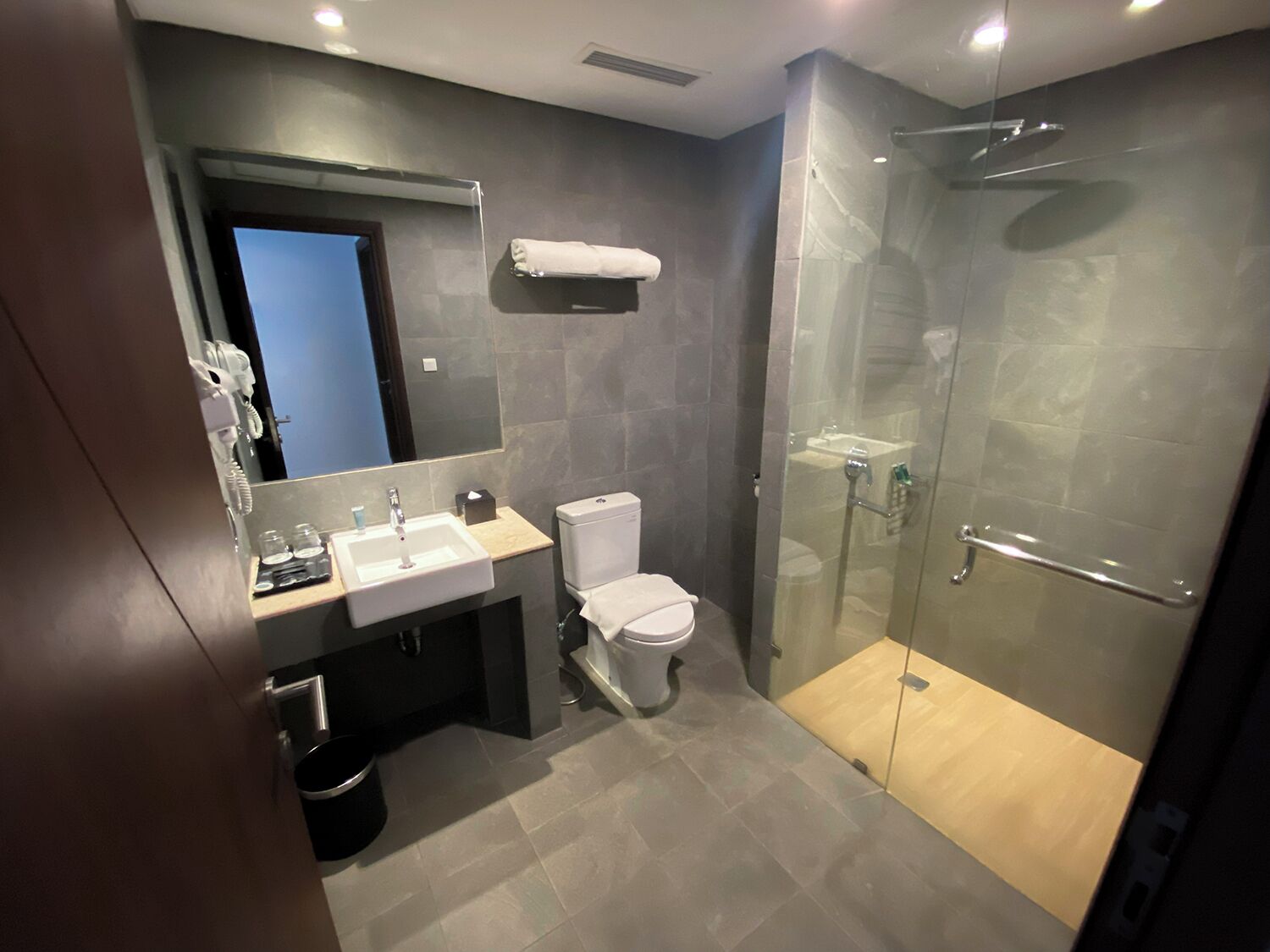 Un baño moderno con ducha envuelta en vidrio, inodoro blanco y lavamanos con espejo superior.
