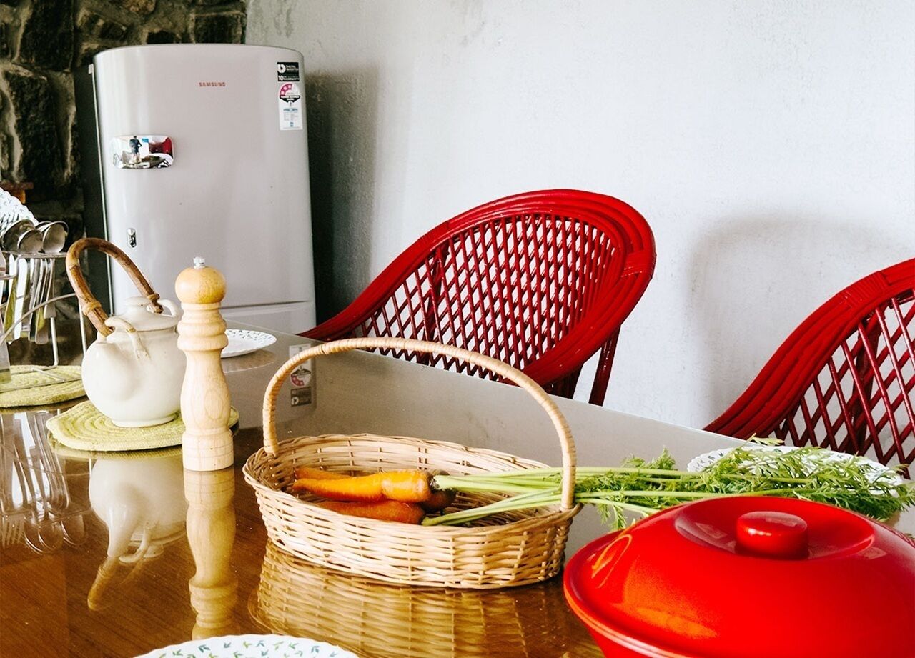 Una mesa de comedor con una cesta de mimbre que contiene verduras, una olla roja y una tetera blanca.