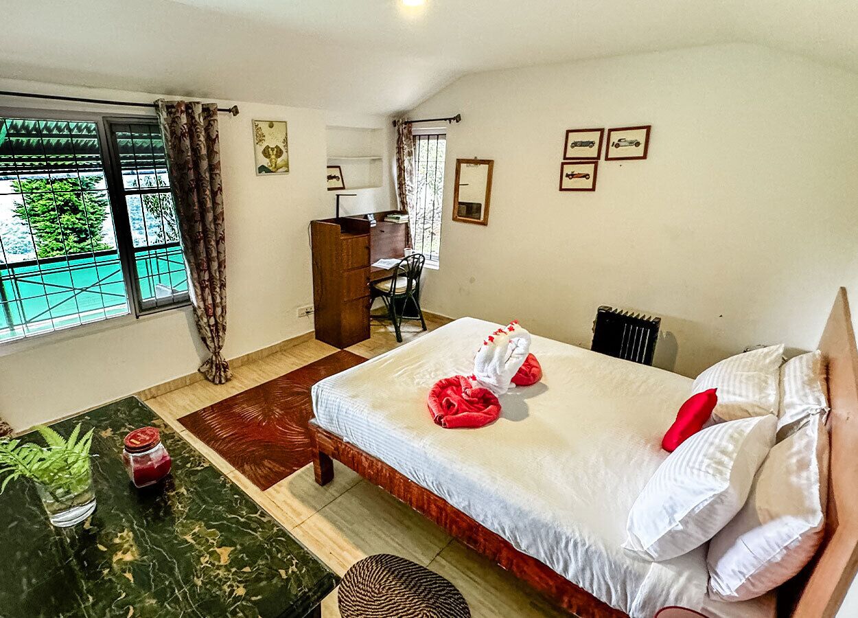 A neatly arranged hotel room with a bed, a desk, a chair, a small table with a plant, and a view of the outdoors through a window.