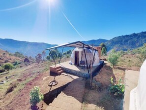 A small, single-story structure with a covered porch and a door, situated on a hillside with a view of distant mountains. - Manas Valley View bungalow  (Kodaikanal)