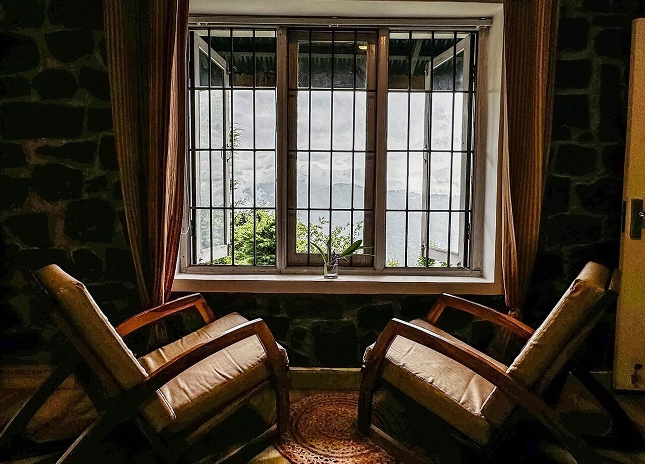 A room with a stone wall, a window with metal bars, two wooden chairs, and a small table with a plant.