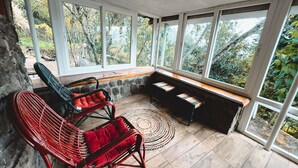 A covered outdoor seating area with wicker chairs and a bench, surrounded by large windows offering a view of trees and a stone wall. - Manas Valley View bungalow  (Kodaikanal)