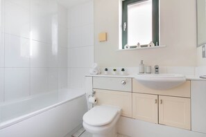 A modern bathroom with a white bathtub, toilet, and sink. There is a small shelf with toiletries and a mirror above the sink. - Cozy 1BR Near Tower Bridge - St Paul's Cathedral (London)