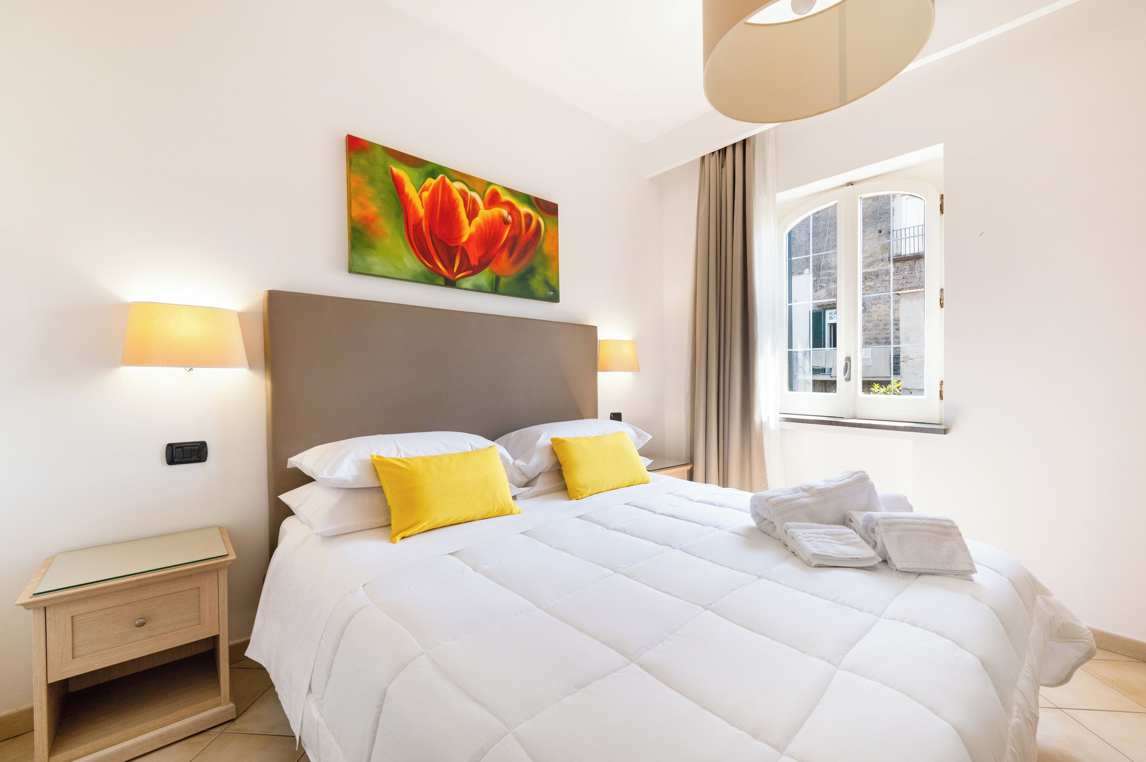Un lit bien fait, avec une literie blanche et deux coussins jaunes, une table de chevet avec une lampe et un tableau représentant une fleur accroché au mur.