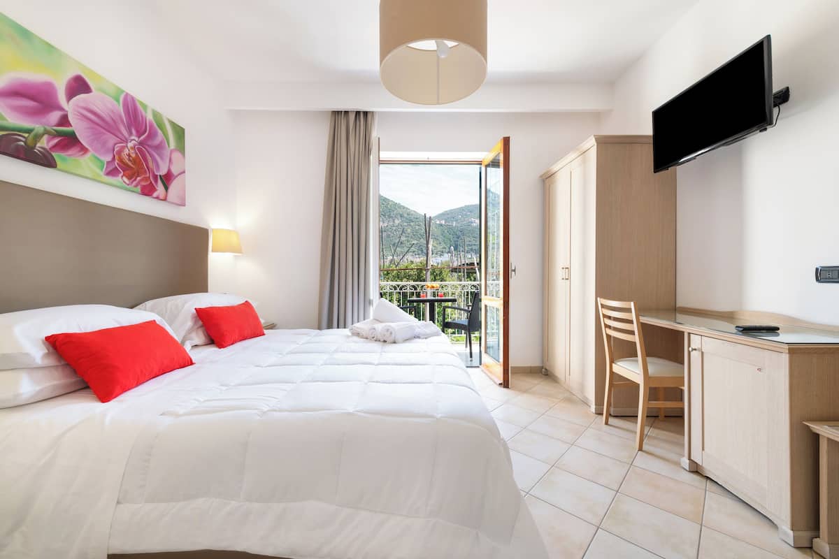 A hotel room with a bed, a desk, a TV, and a balcony with a view of the mountains.