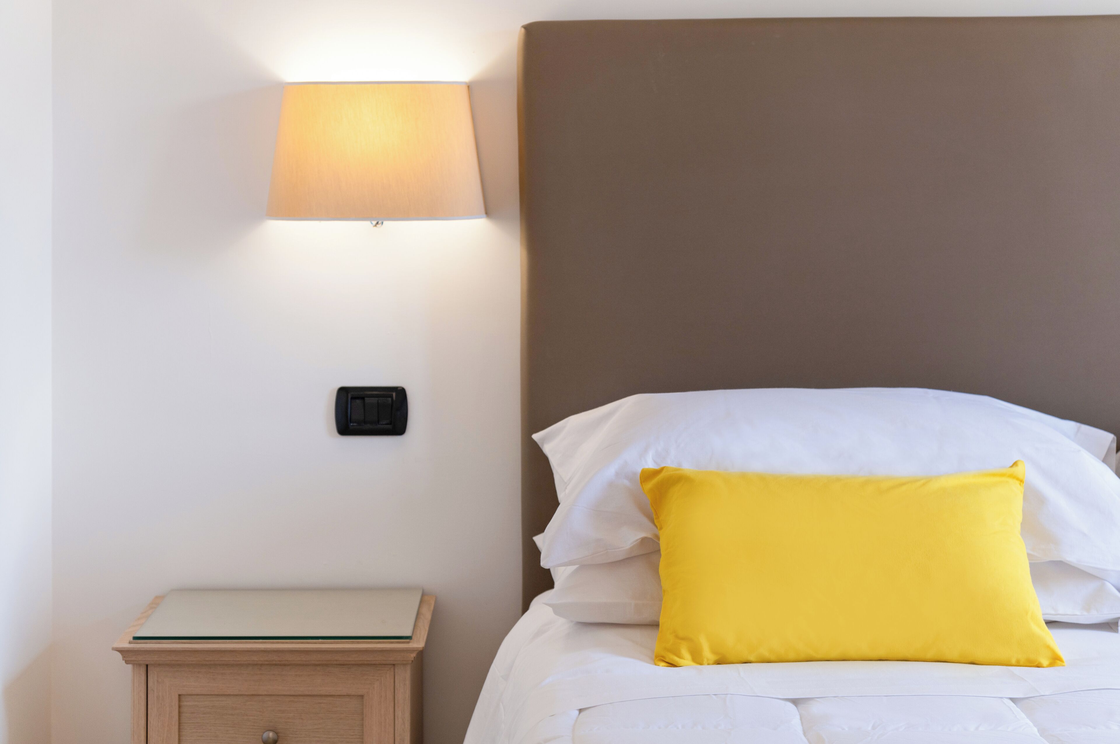 Un lit bien fait, avec des oreillers blancs et jaunes, une lampe de chevet et une petite table de nuit en bois sur laquelle repose un livre.